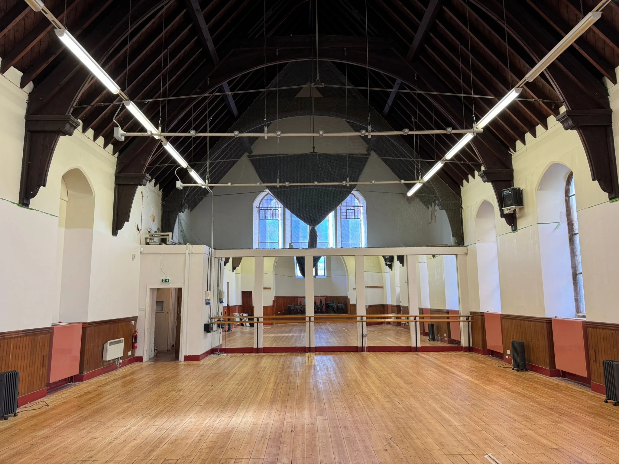 The North Hall viewed from the stage end, showing the full length of the hall with polished wooden floors, a vaulted timber ceiling, arched stained glass windows, and a mezzanine balcony