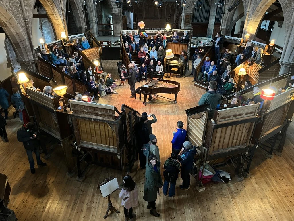 A Pianodrome community event in the South Hall — people gathered around a grand piano at the centre, surrounded by a circular arrangement of upright pianos, with the hall's original stone arches above